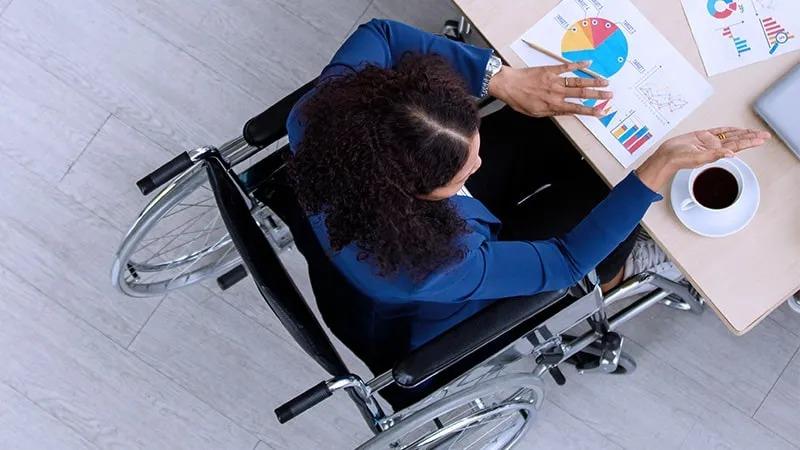 Person in a wheelchair working at a desk with charts and graphs, gesturing toward a coffee cup, viewed from above.