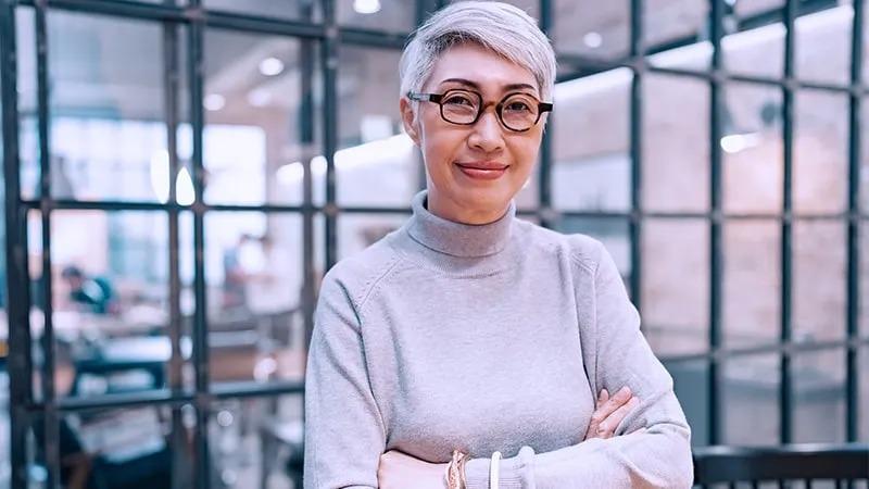 Confident older woman with short gray hair and glasses, wearing a light gray turtleneck sweater, standing with arms crossed in a modern office space.