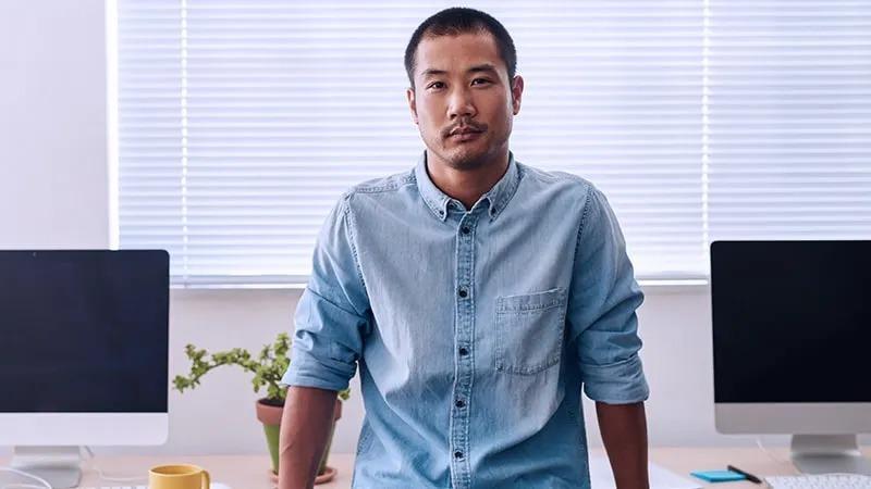 Man in a denim shirt sitting on the edge of a desk in an office, with two computer monitors, a yellow mug, and a potted plant in the background.