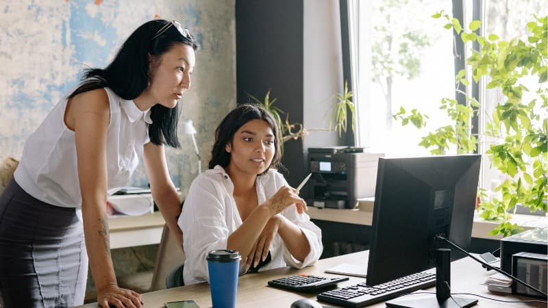 Two women in an office setting collaborating at a desk, with one seated and pointing at a computer screen while the other stands beside her, both focused on the discussion.