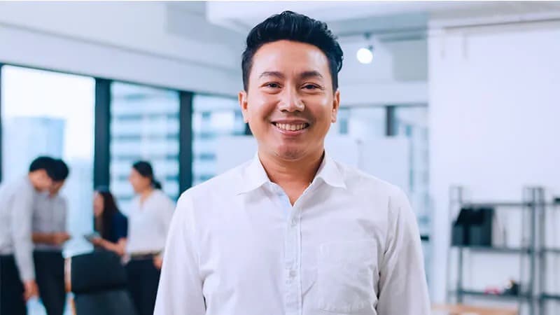 Smiling man in a white shirt standing in an office environment, with colleagues collaborating in the blurred background.
