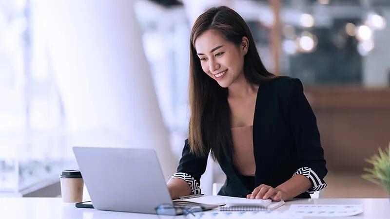 Smiling woman in business attire working on a laptop at a desk, with a coffee cup and notebook beside her in a bright modern office.