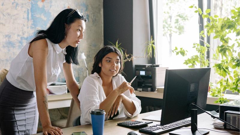 Two women in an office setting collaborating at a desk, with one seated and pointing at a computer screen while the other stands beside her, both focused on the discussion.