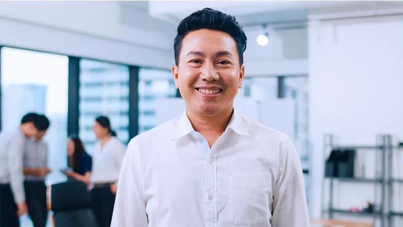Smiling man in a white shirt standing in an office environment, with colleagues collaborating in the blurred background.