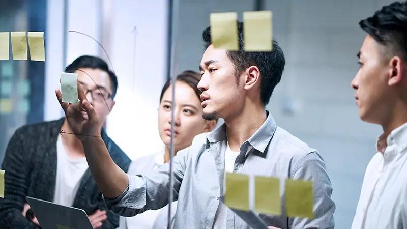 Group of young professionals collaborating in a meeting, with one man placing sticky notes on a glass wall while others watch attentively.